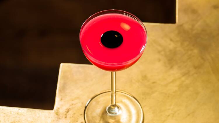 A bright pink cocktail in a martini glass with a black circle floating in its centre, sitting on a bronze table with a zig-zag edge