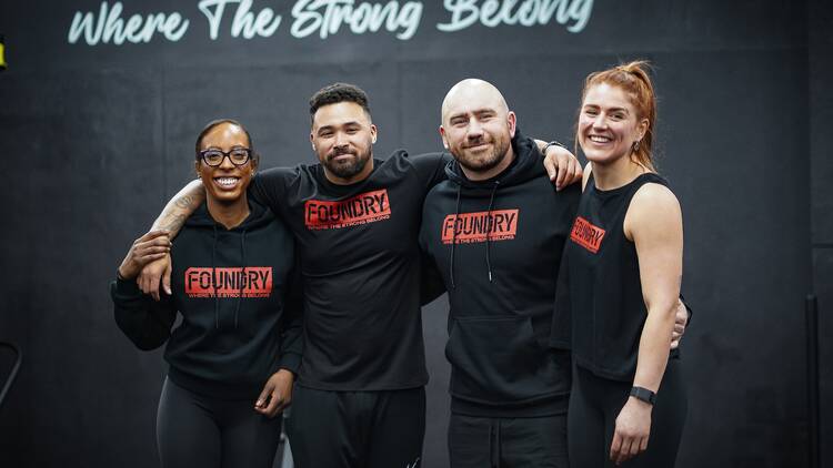 Four trainers dressed in black tops with an orange Foundry logo on them huddles together and smiling