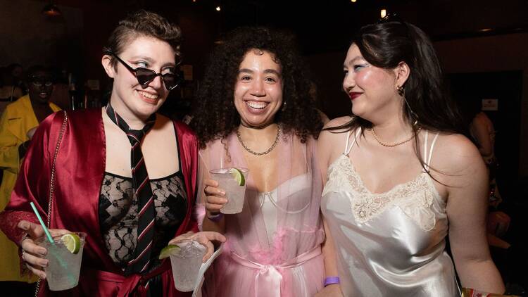 Three people in lingerie-inspired outfits pose while holding drinks in their hands.