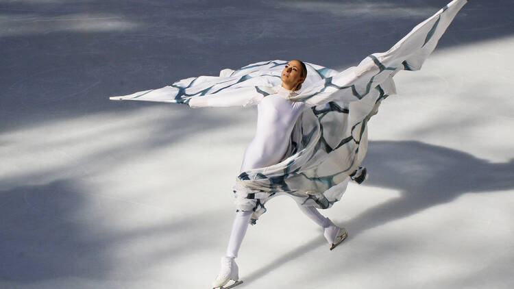 An ice dancer glides across the rink in a white outfit with a blue-and-white cape.