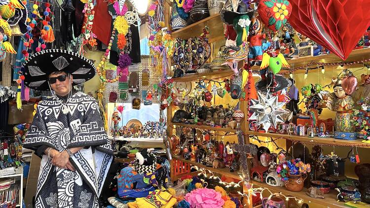 A man in a traditional Mexican outfit stands inside of a store filled with colorful Mexican folk art.