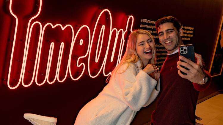 Two people taking a selfie in front of the Time Out sign at Time Out Market Bahrain