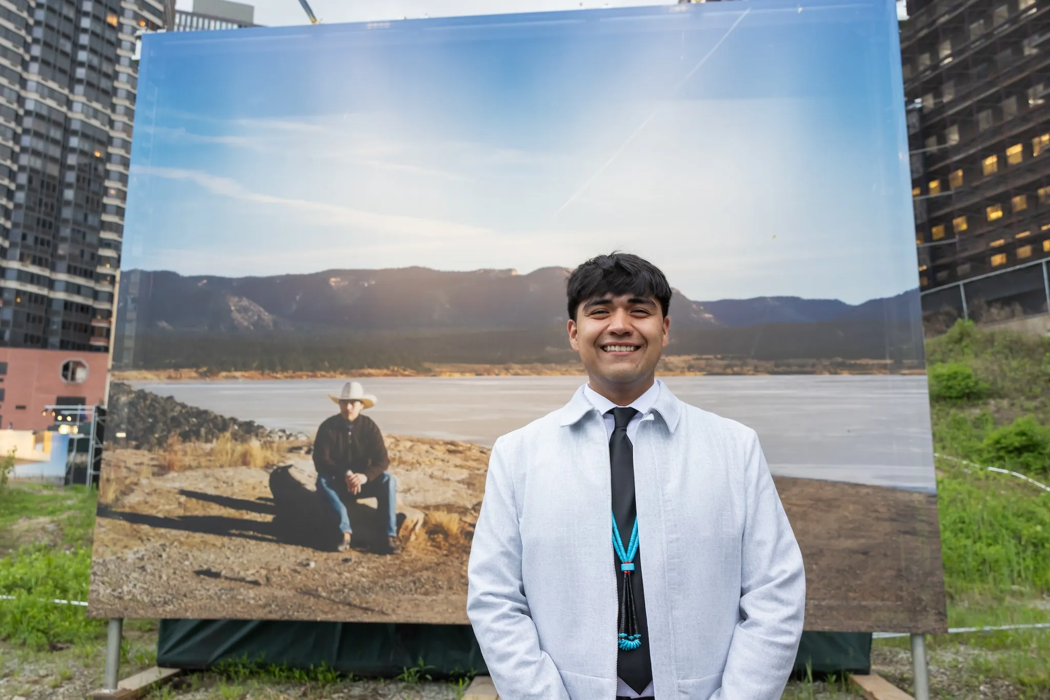 A man wearing a black tie with turquoise beads and a gray jacket stands in front of a large photograph at an art exhibit.