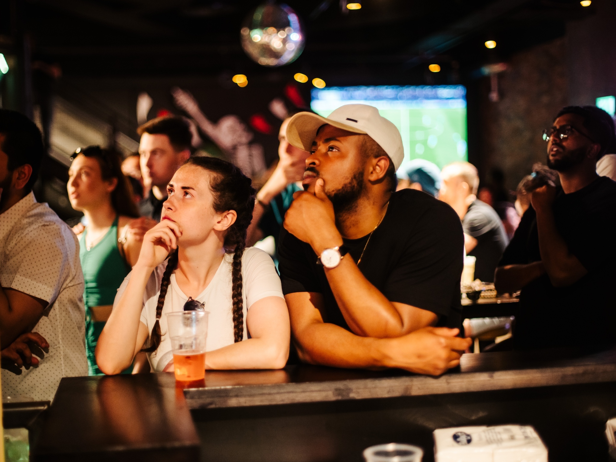 A man and a woman stare up at a screen showing a football match, at Bat & Ball Covent Garden