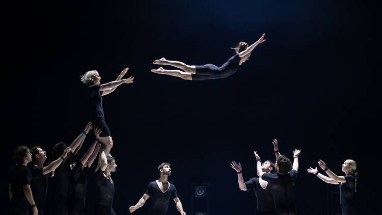 A circus performer flying through the air while other acrobats stand ready to catch.
