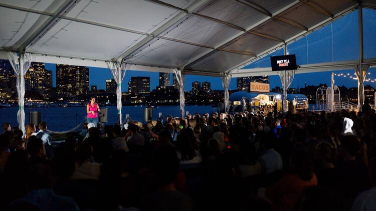 comedian performing in front of an audience with Boston in background at night