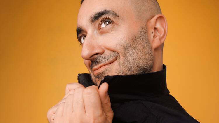 A man poses for a headshot in a black jacket against an orange background. A man poses for a headshot in a black jacket against an orange background.