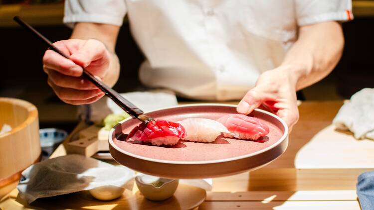 A chef in white serves nigiri from a pink plate with chopsticks