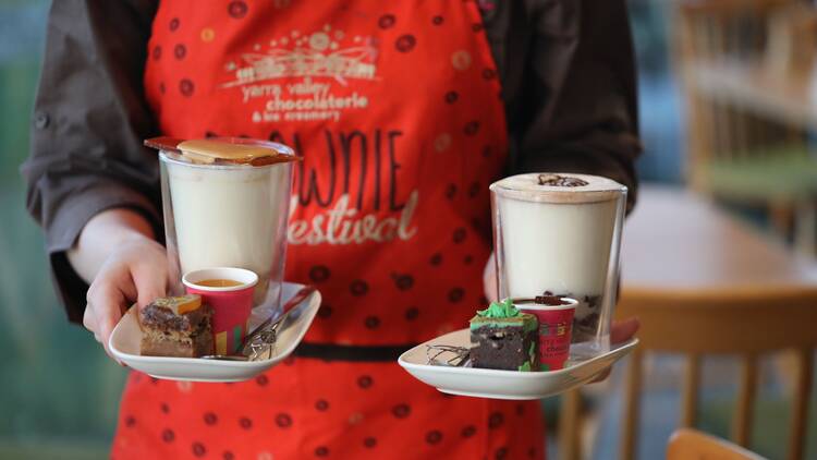 A person in a red apron holding plates with hot chocolates and brownies.