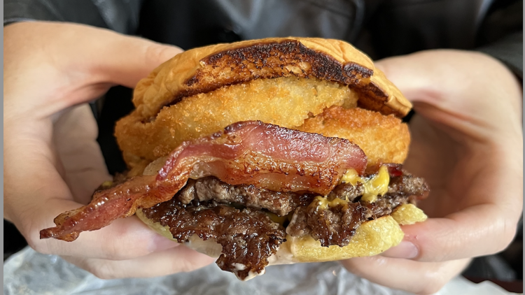 Casa 13urger A person holding a burger with bacon, cheese and an onion ring