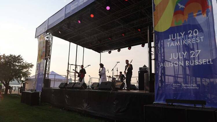 Musicians perform on a stage with a pink sunset in the background.