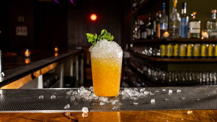 An orange cocktail with pebble ice and spring of mint sitting on a bar