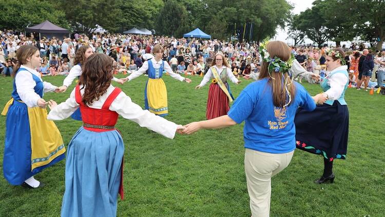 A group of women hold hands while dancing in a circle. Some wear traditional Swedish dress.