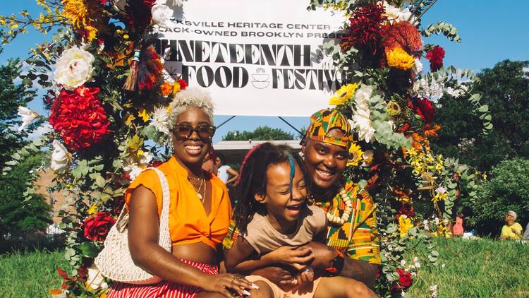 A group of three people sitting in front of a sign with flowers that reads "Juneteenth Food Festival"