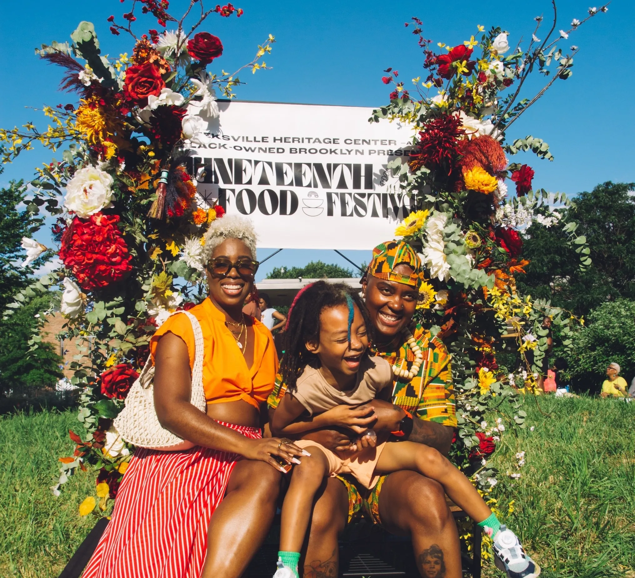 A group of three people sitting in front of a sign with flowers that reads "Juneteenth Food Festival"
