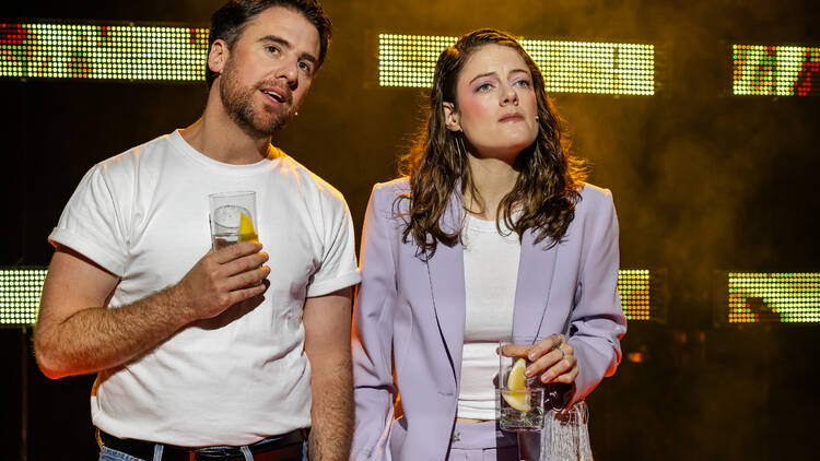 Two actors in 'Heartbreak Hotel' stand on stage holding drinks.