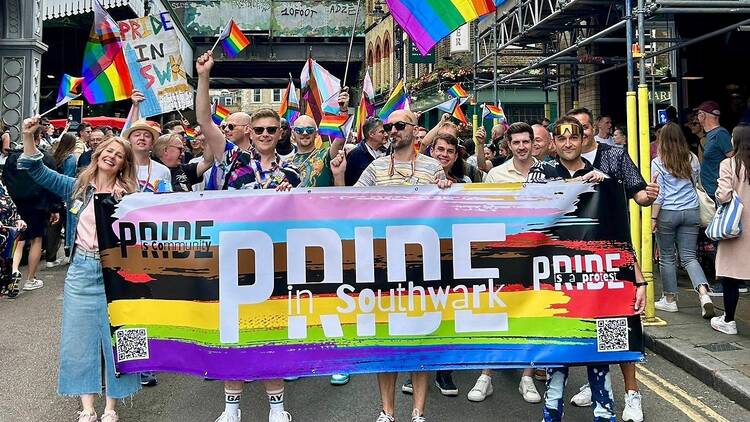 Pride in Southwark attendees march behind a Pride flag banner Pride in Southwark attendees march behind a Pride flag banner