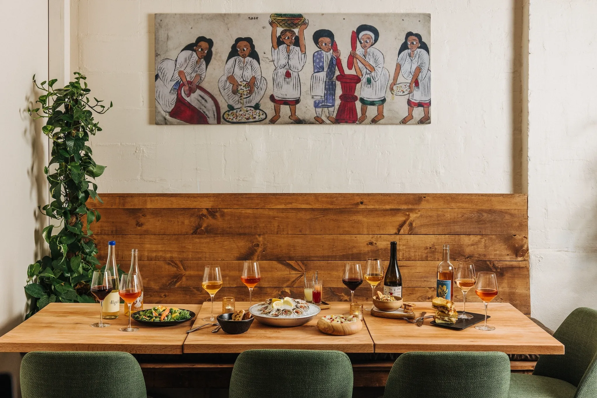 A wooden booth with green chairs. Above it an art piece depicting women preparing food.