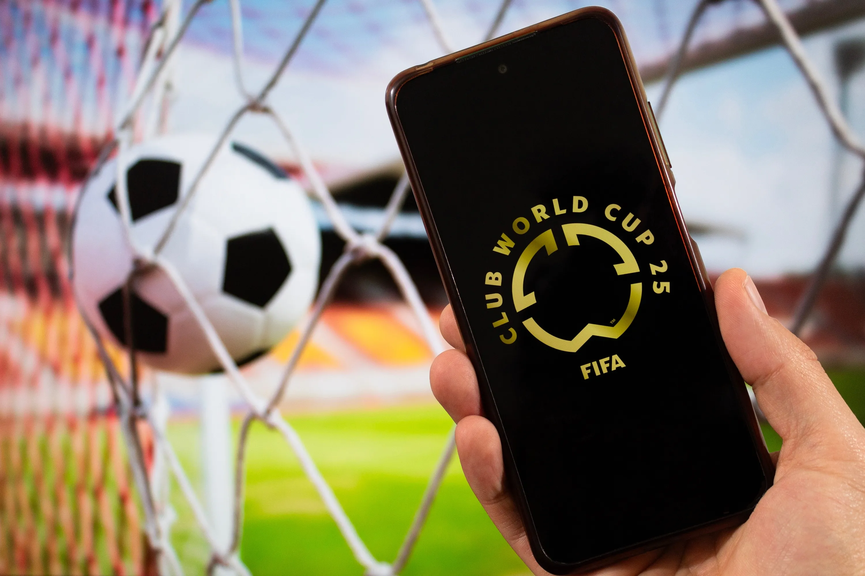 A male hand holding a smartphone with fifa club world cup golden logo and blurred football ball at background