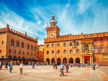 Italy Piazza Maggiore in Bologna old town tower of hall with big clock and blue sky on background. Antique buildings terracotta galleries Italy Piazza Maggiore in Bologna old town tower of hall with big clock and blue sky on background. Antique buildings terracotta galleries