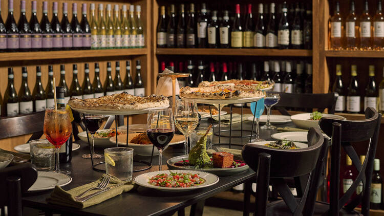 Pizzas and drinks on a table with stacked wine bottles on the wall