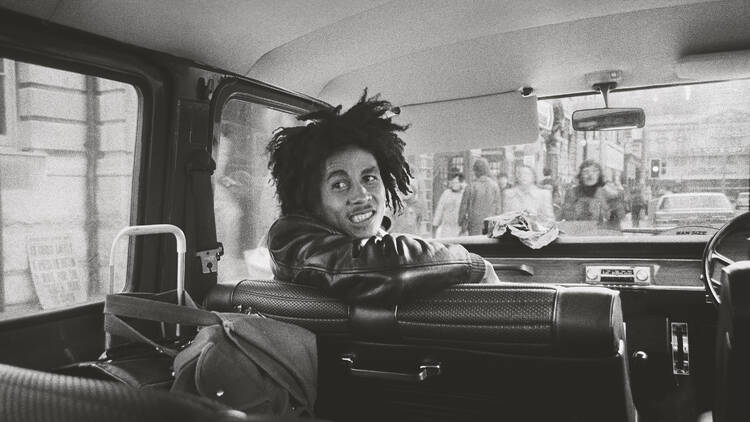 Black and white photograph of Bob Marley in the passenger seat of a van with his body partially turned towards the back of the vehicle.