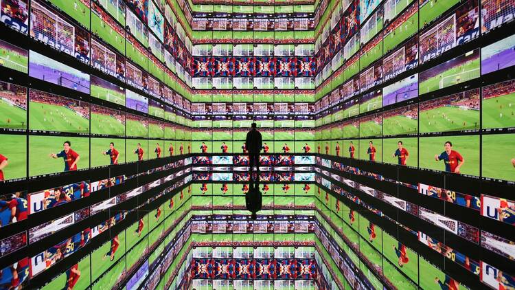 Bright screens on three walls show a soccer player on field; a person stands in the middle watching.