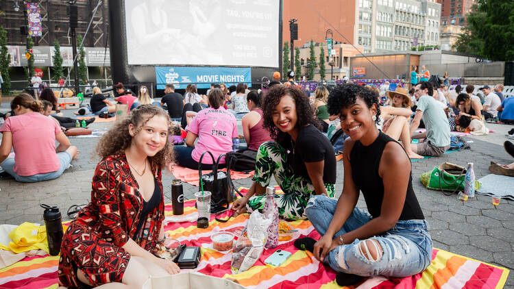 A group of young women sit on a picnic blanket watching a movie.