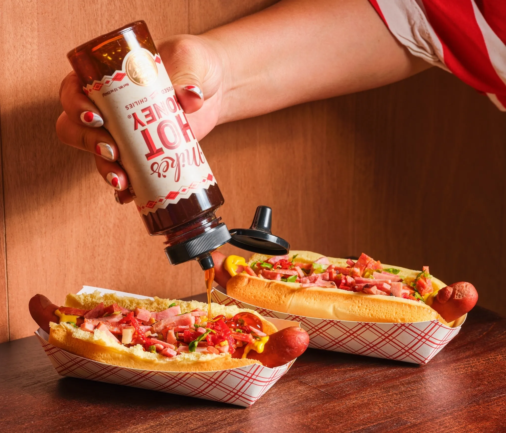 two hot dogs with cold cuts and a person drizzling Mike's Hot Honey on top