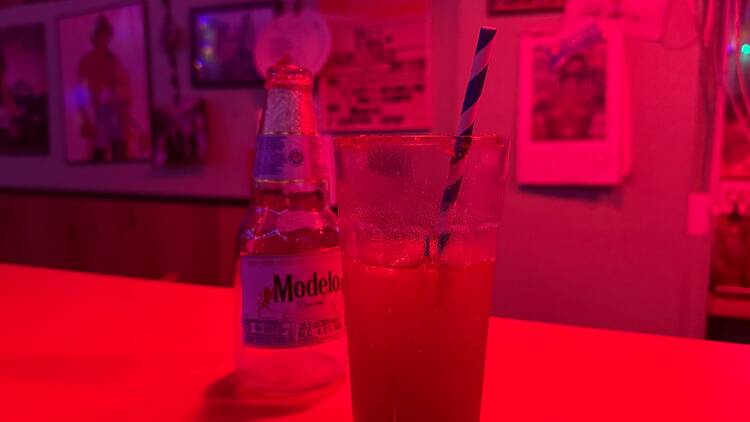 A glass containing a stripy paper straw and a half drunk michelada stands next to a bottle of Modelo on a bend bathed in red light