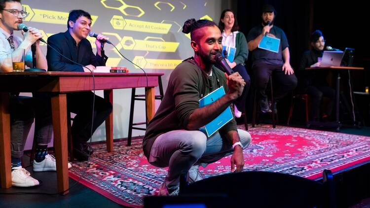A group of comedians on a stage with one in the center crouching over a red patterned rug spelling a word and making contestants in the background laugh