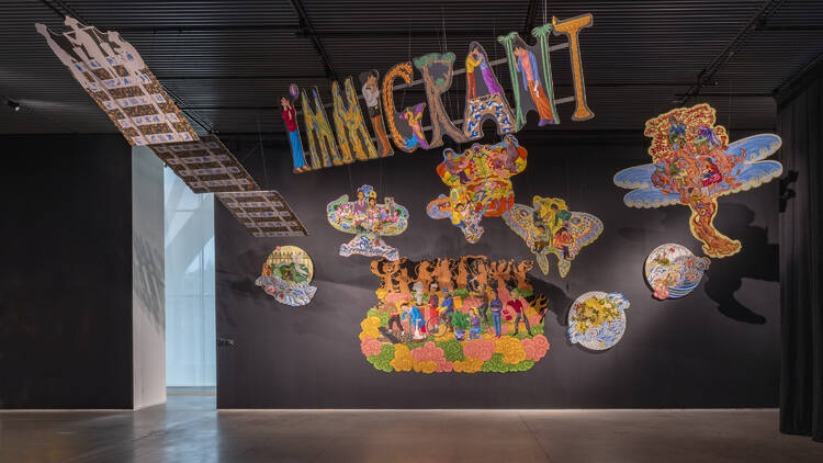 Several multi-colored art pieces hanging from the ceiling and the wall, the main one saying "Immigrant" 