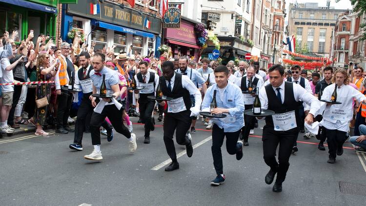 A line of waiters dressed in their uniforms race through the streets of Soho past the French House pub, carrying trays loaded with champagne bottles and ashtrays