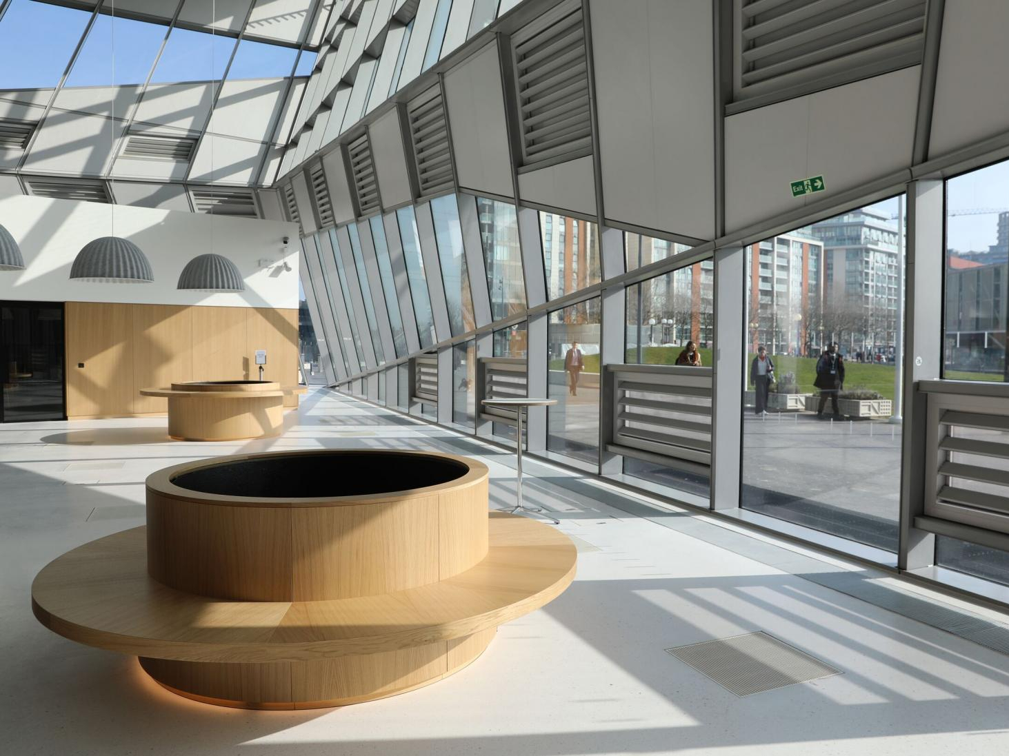 Interior of City Hall, with a large circular wooden bank of seating in the foreground, a glass roof and windows looking out onto a green lawn