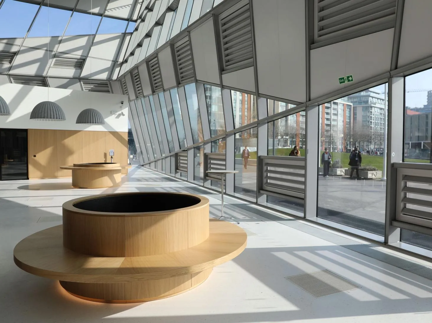 Interior of City Hall, with a large circular wooden bank of seating in the foreground, a glass roof and windows looking out onto a green lawn