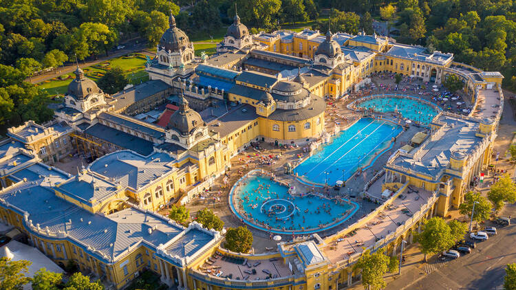 széchenyi baths. Budapest filmed from a drone. We see a large swimming pool and a beautiful building. People swim in the pool. Around the building there is a Park with trees. Sunny summer day.