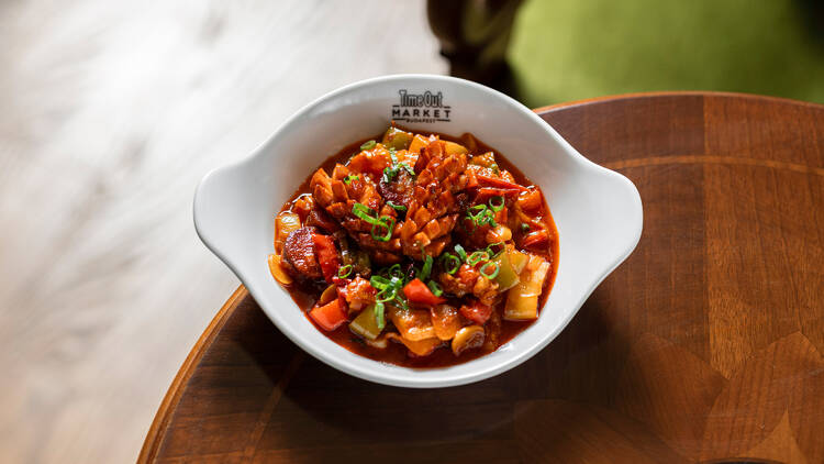 A close-up of the traditional Hungarian stew (lecsó) served in a branded Time Out Market Budapest bowl, garnished with green onions and presented on a carved wooden table.