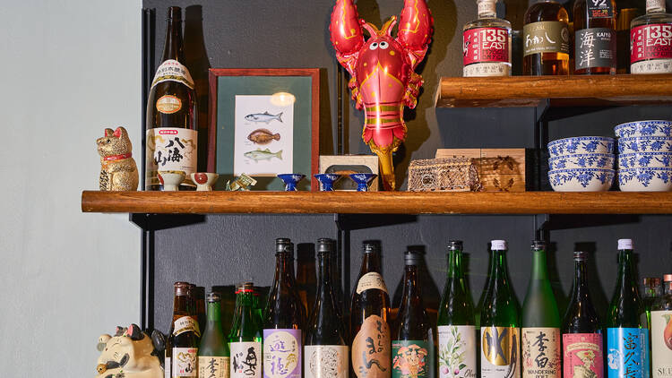 Three shelvves on the wall hold a variety of Japanese spirits along with accoutrements like cats and lobsters