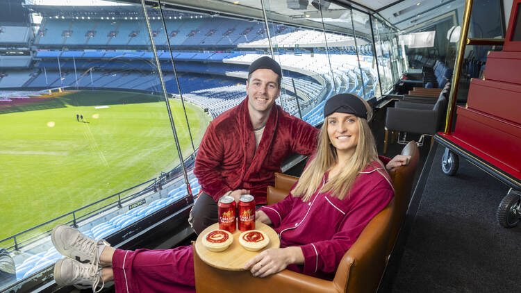 Two people in pjyamas sit in comfy chairs overlooking the MCG.