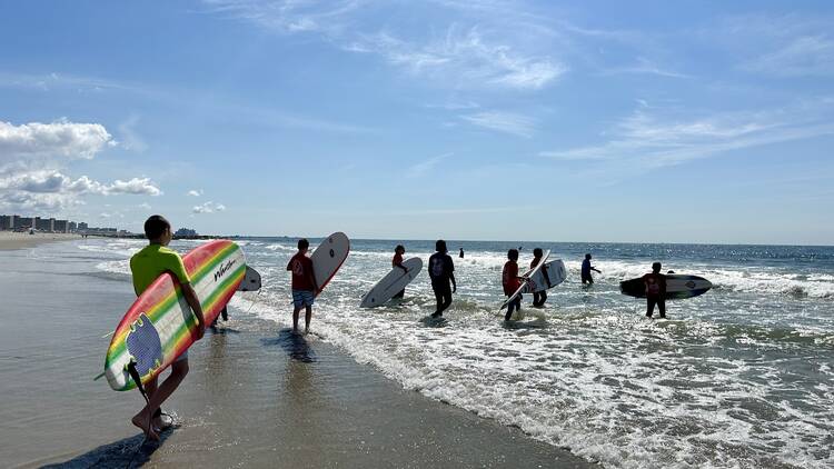 A group of kids carries surfboards in the ocean with Black Surfing Rockaway.