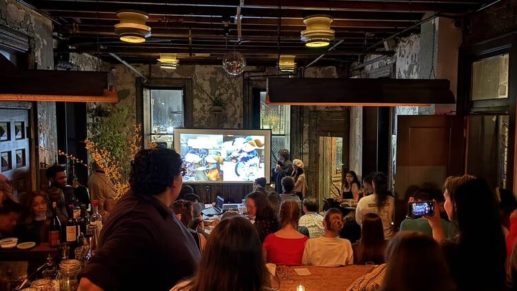 A crowd gathered in a bar in front of a screen which displays a slide deck