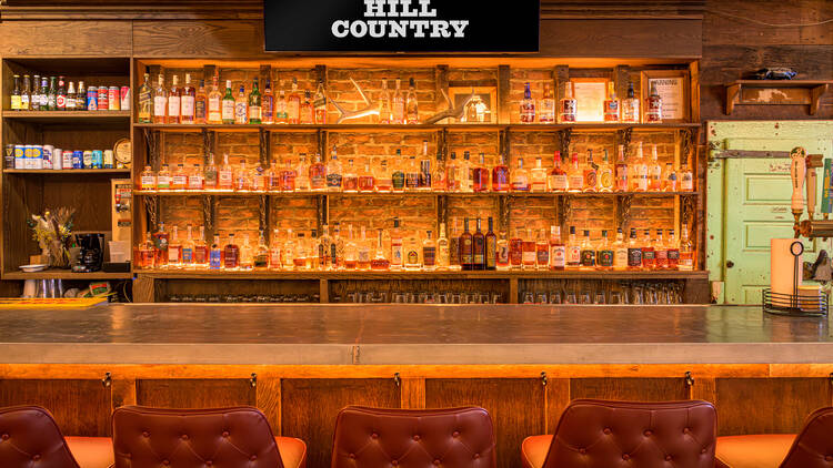 An illuminated bar with red stools. Above it reads a sign that says Hill Country