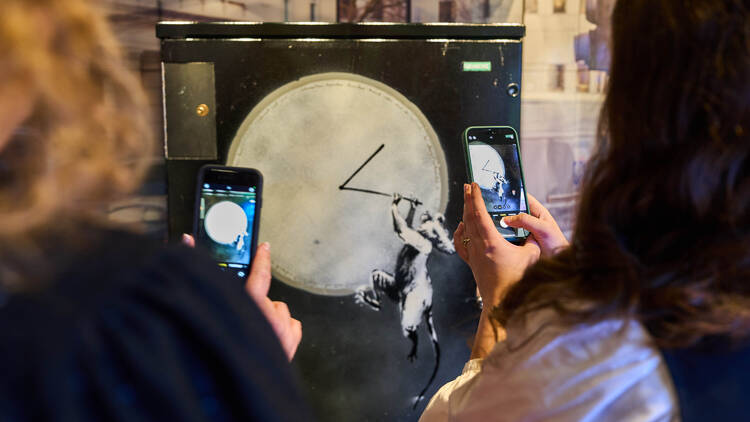 Two people taking a photo of the Banksy on display in the London Transport Museum
