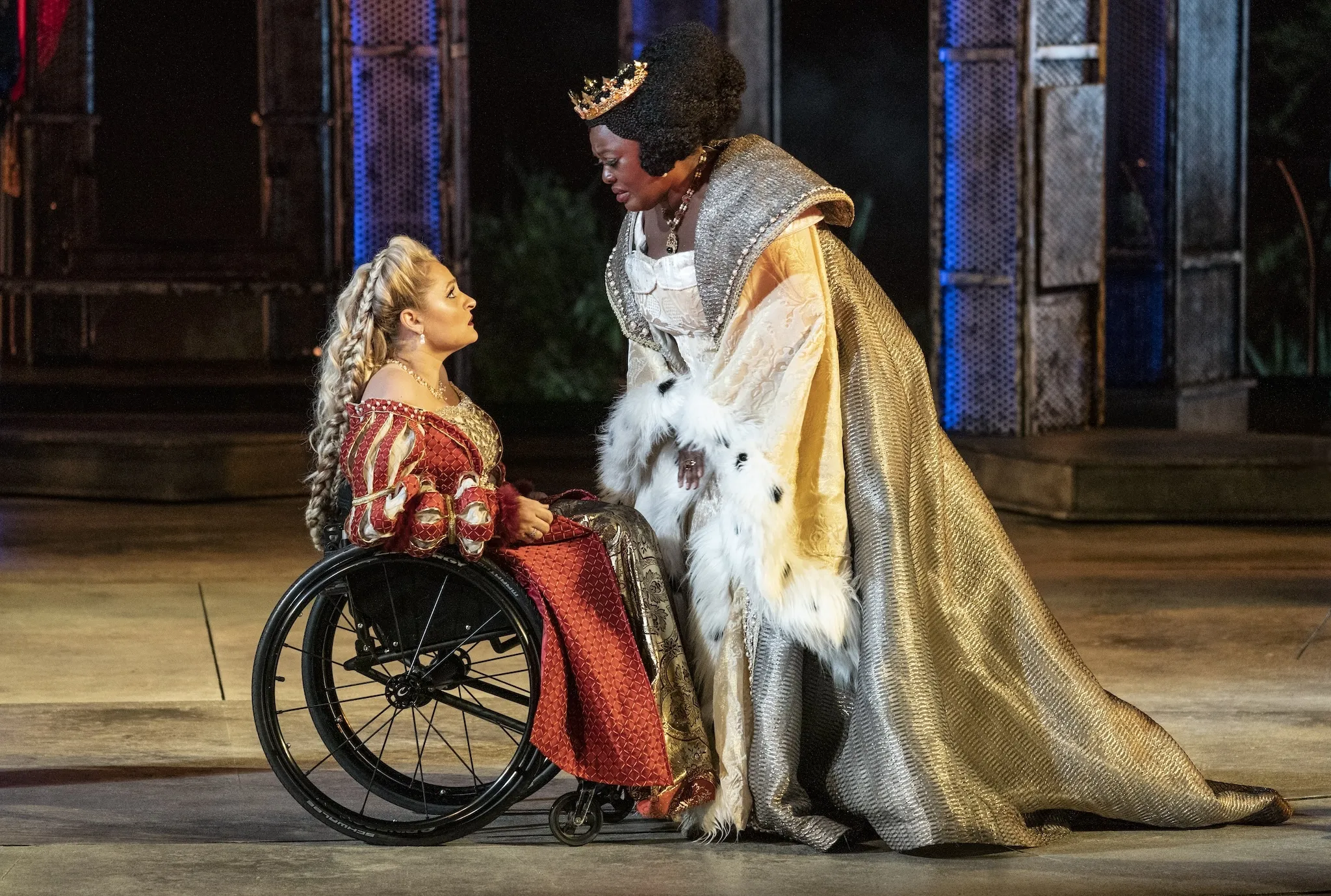 Two actresses on stage. The actress on the left wears a red dress and uses a wheelchair. On the right, the actress is in a white-and-gold dress standing next to her co-star.