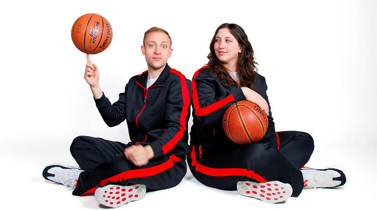 Two people dressed in black and red tracksuits and holding basketballs sit against a white backdrop. Two people dressed in black and red tracksuits and holding basketballs sit against a white backdrop.