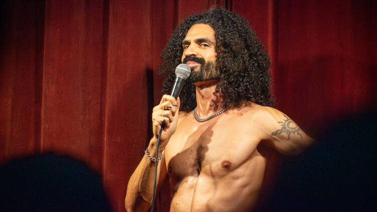 A man, seen from the waist up, performs comedy in the nude.