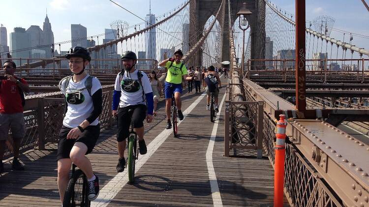 Unicyclists ride in a straight line on a bridge with the cityscape in the background