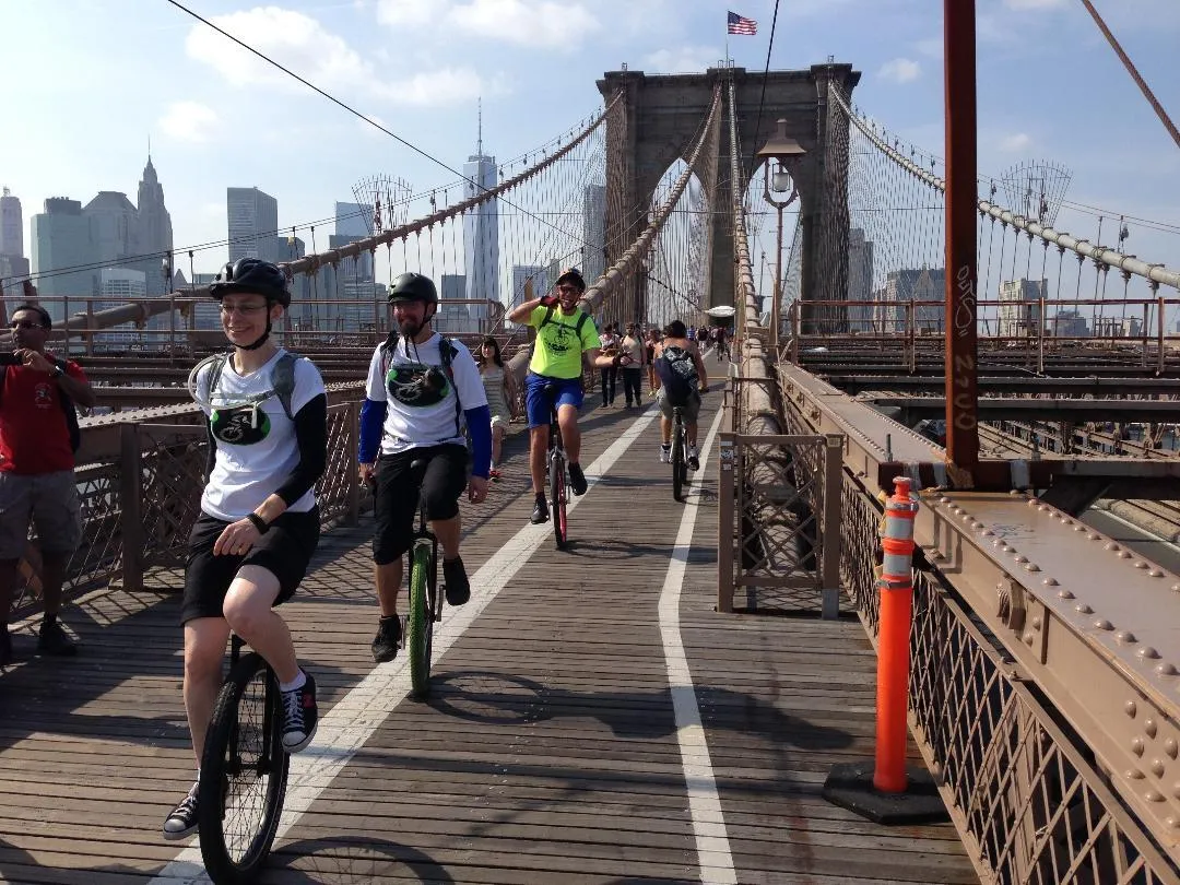 Unicyclists ride in a straight line on a bridge with the cityscape in the background