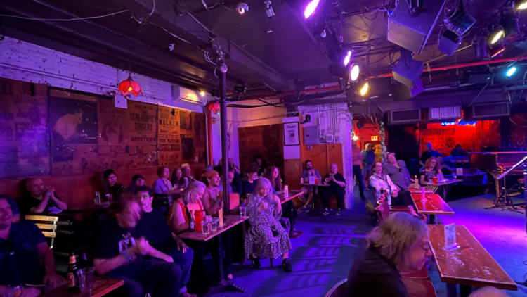 A room lit in purple lighting with many people sitting at tables, looking up at a stage. There are posters on the wall and a microphone on the stage for readers.
