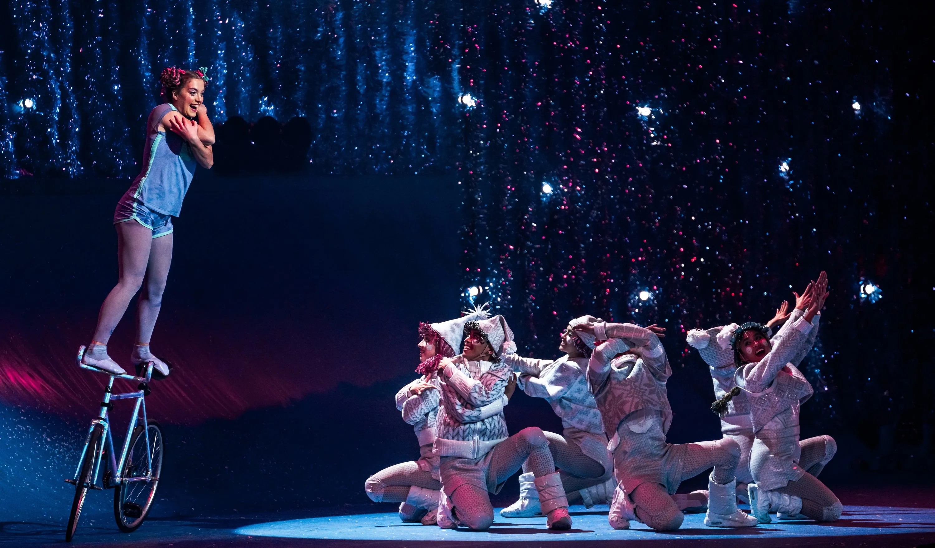 A girl balancing on a bike in front of a group of performers in grey show suits on the ground
